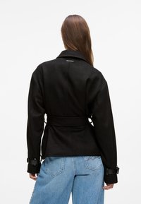 Czarna kurtka z mieszanki wełny z paskiem w talii, szerokimi klapami i czarnymi akcentami metalowymi. Noszona na denim o jasnym odcieniu z wyhaftowanymi detalami.