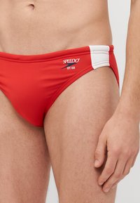 Røde svømmebokser lavet af strækbart materiale, med hvide sidepaneler og Speedo-logoet. Designet har en tætsiddende, lavtaljet form.