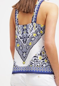 Top sin mangas con base blanca, que presenta un patrón floral azul y amarillo, decorado con detalles intrincados y un ribete en contraste.