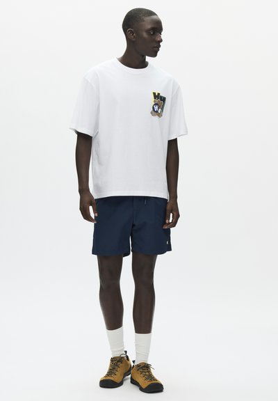 Νέος άνδρας που φοράει λευκό T-shirt με γραφικό σχέδιο, μπλε σκούρες βερμούδες, λευκές κάλτσες και καφέ παπούτσια πεζοπορίας, στέκεται μπροστά σε λευκό, απλό φόντο.