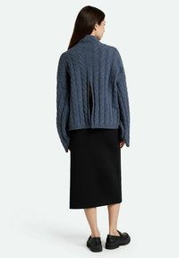 Cardigan en tricot épais bleu foncé avec un col haut, un dos ouvert et des manches surdimensionnées, associé à une jupe midi noire et des chaussures noires.