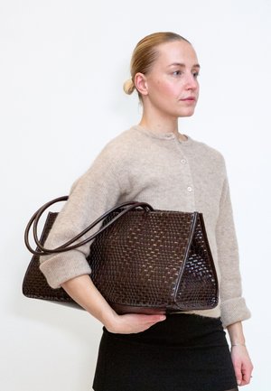 Femme aux cheveux blonds attachés en chignon porte un pull beige et une jupe noire, tenant un grand sac à main tressé marron foncé sous le bras.