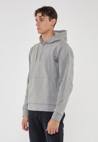 Felpa grigia con cappuccio e tasca a marsupio, logo ricamato, polsini a costine e tessuto morbido, indossata con jeans scuri.
