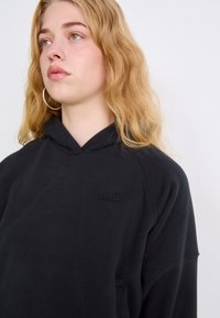 Sudadera con capucha negra de corte relajado, que presenta un logo texturizado en el pecho y una tela suave. El modelo tiene el cabello rizado y claro.