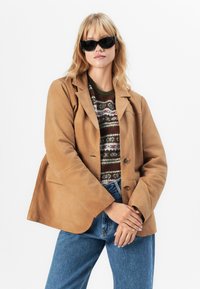 Kvinne som har på seg svarte solbriller, en beige suedjakke over en mønstret genser, og blå jeans, poserer med armene krysset mot en hvit bakgrunn.