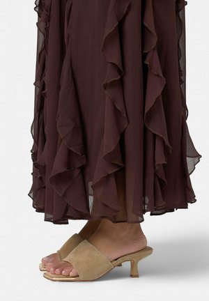 Pies calzando mocasines mule de tacón bajo de ante beige con una falda midi marrón oscura con volantes sobre un fondo blanco.