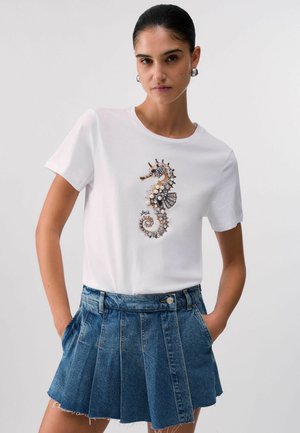 Donna che indossa una t-shirt bianca con un motivo di cavalluccio marino gioiellato e una gonna di jeans blu plissettata con le mani nelle tasche.