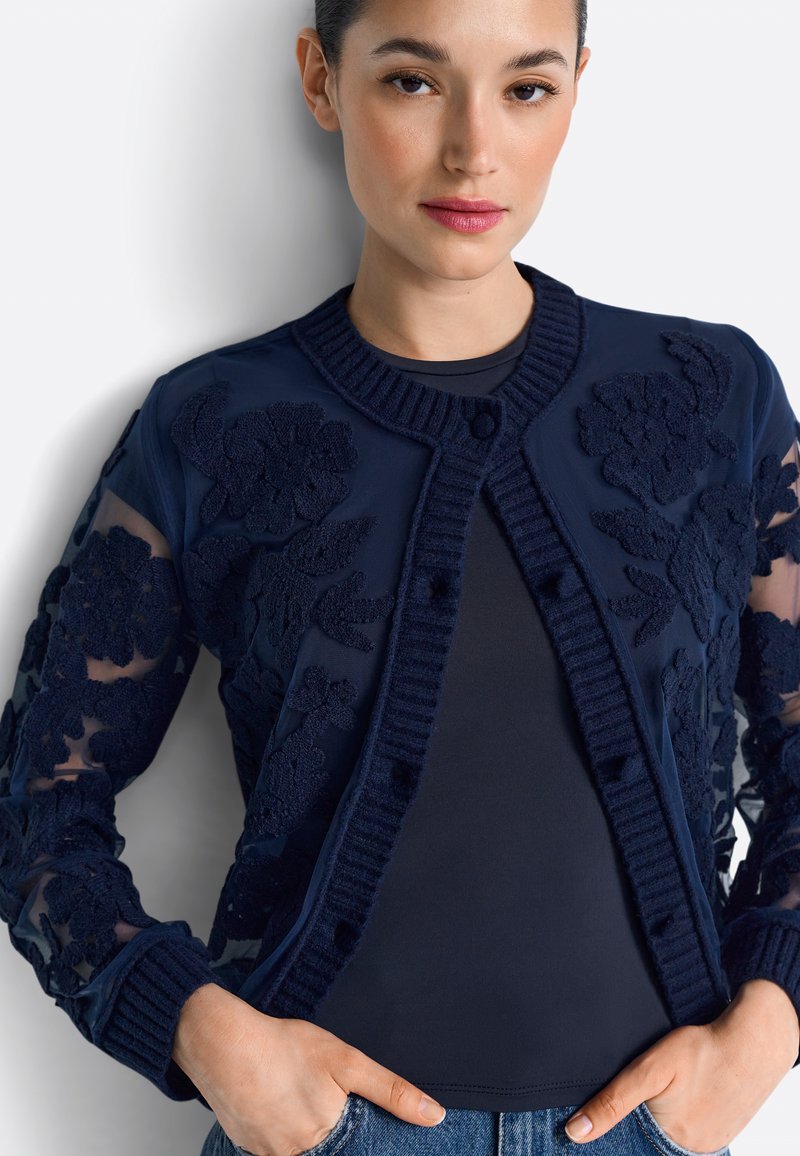 Cardigan blu navy con ricami floreali, maniche trasparenti, collo a coste e chiusura frontale con bottoni; abbinato a una maglia scura e denim.