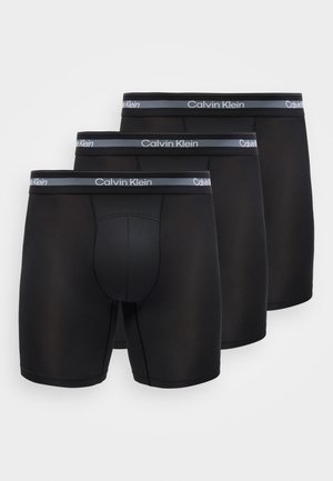 Trois boxers Calvin Klein noirs empilés, avec une ceinture grise portant le logo Calvin Klein en blanc.