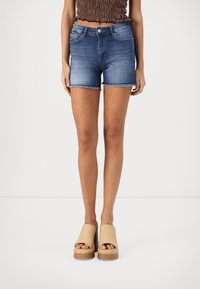 Pantalones cortos de mezclilla en azul oscuro, de tiro medio y con dobladillo deshilachado. Combinados con sandalias de plataforma beige que cuentan con tacones de madera y diseño de punta descubierta.