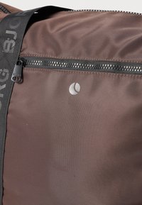 Καφέ υφασμάτινη τσάντα με οριζόντιο φερμουάρ, λογότυπο σε ασημί χρώμα και μαύρη ρυθμιζόμενη ζώνη με ανάγλυφα γράμματα για επιπλέον λεπτομέρεια.