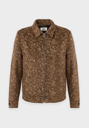 Homme portant une veste marron à imprimé léopard, une chemise bleu clair, un pantalon beige et des chaussures marron, debout devant un fond uni clair.