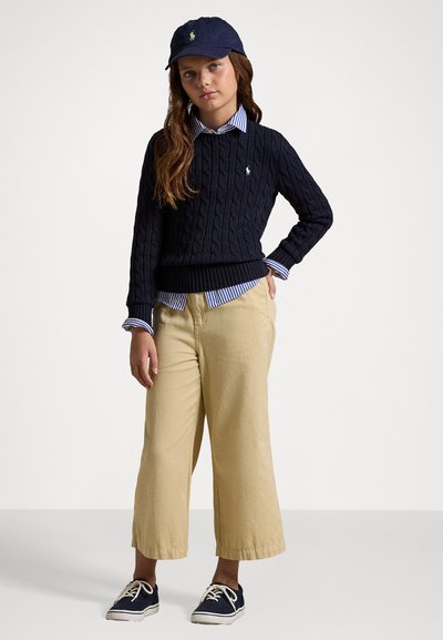 Suéter azul-marinho de malha trançada, camisa colarinho listrada em azul e branco por baixo, calças largas bege e ténis azul-marinho com atacadores brancos.