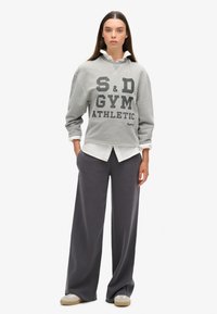 Grijze sweatshirt met vetgedrukte zwarte tekst "S&D GYM ATHLETIC," gelaagd over een witte blouse, gecombineerd met brede grijze broek en beige schoenen.