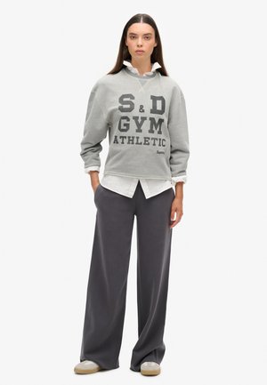 Felpa grigia con bold nero "S&D GYM ATHLETIC," sovrapposta a una camicia bianca con colletto, abbinata a pantaloni grigi larghi e scarpe beige.