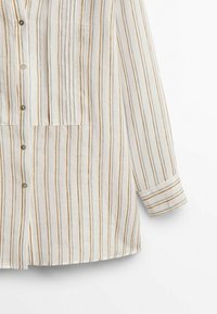 Chemise blanche à manches longues avec des rayures verticales orange et marron, boutonnée sur le devant et panneau décoratif cousu sur la poitrine.