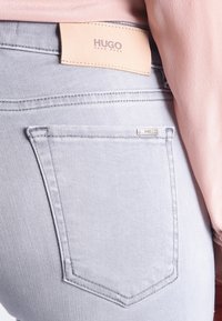 Jean en denim gris avec une étiquette en cuir HUGO à la taille, comportant des poches arrière et des détails de couture subtils. Texture lisse.