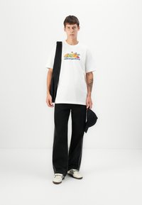 T-shirt bianca con grafica colorata "Adidas Originals", abbinata a jeans neri a gamba larga con strisce laterali e sneakers bianche. Borsa nera e cappellino.