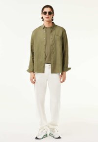 Uomo che indossa una camicia verde oliva sopra una t-shirt abbinata, pantaloni bianchi, sneakers bianche e occhiali da sole scuri, in piedi contro uno sfondo bianco.