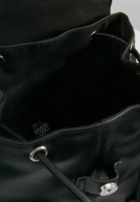 Černý stahovací pytel s měkkou texturou, kovovými očky a stříbrným uzávěrem na patent. Vnitřek obsahuje textilní podšívku a štítek s logem.