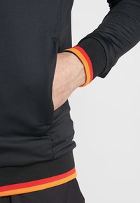 Person som bär en svart jacka med röd och orange rand på ärmarnas och nederdelens fållar, hand placerad i framfickan mot en ljus bakgrund.