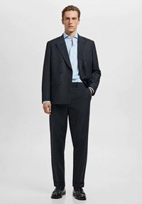 Čierny dvojradový sako cez svetlomodrú košeľu, v páre s tmavými nohavicami a čiernymi topánkami, vytvára formálny, na mieru ušitý vzhľad.