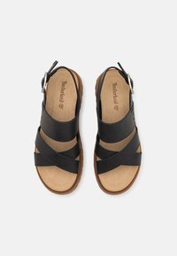 Musta nahkakengät, joissa on ristikkäiset hihnat ja solki, varustettu vaaleanruskealla pehmustetulla pohjalla ja teksturoitulla kumipohjalla.