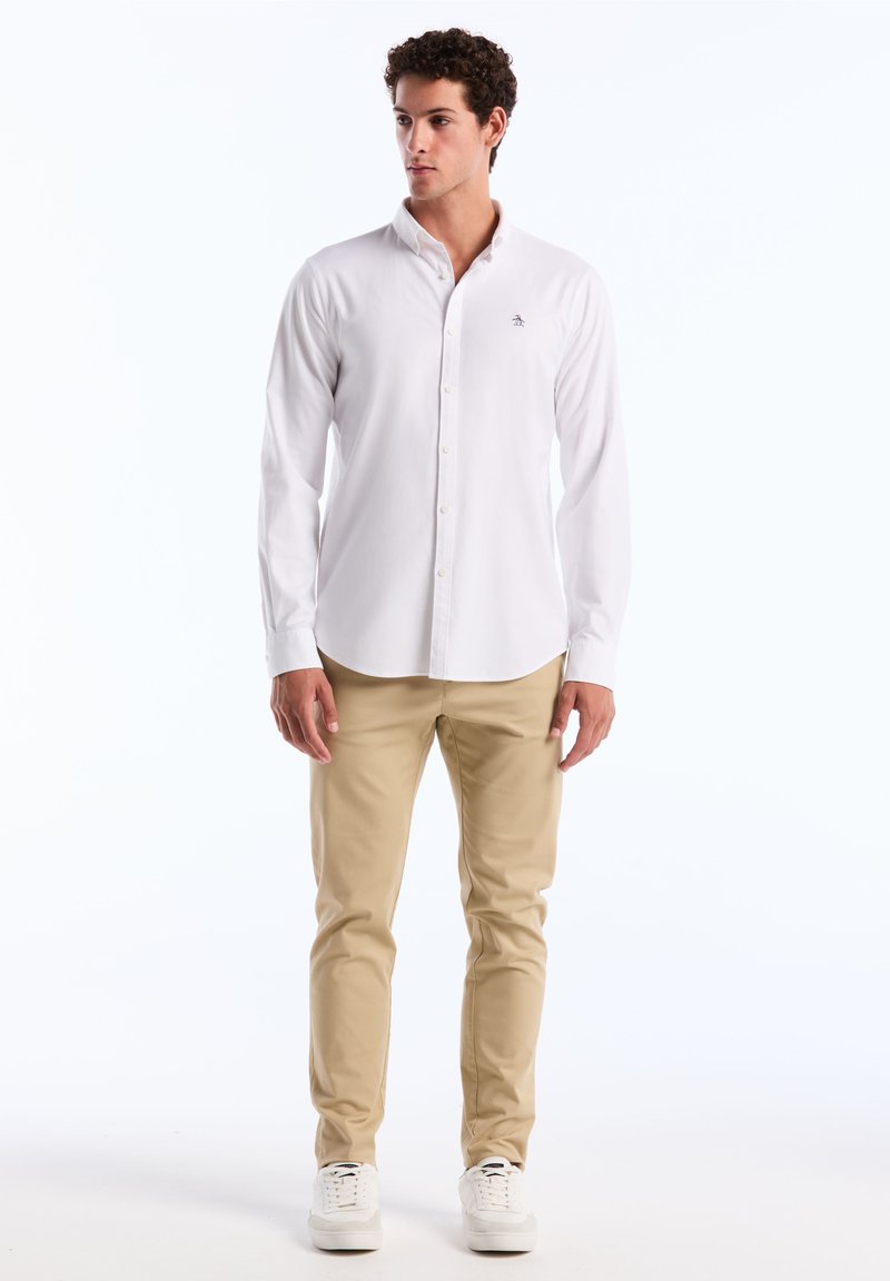 Witte lange mouwen overhemd met knopen en een kraag, voorzien van een klein logo op de borst, gecombineerd met beige broek en witte sneakers.