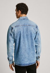 Chaqueta de mezclilla azul clara con puños abotonados y un cuello clásico. Presenta un aspecto desgastado con detalles de costura a lo largo de las costuras.