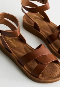 Sandales en cuir marron avec de larges lanières croisées et des boucles pour les chevilles. Dotées d'une semelle intérieure texturée et rembourrée ainsi que d'une semelle plateforme.