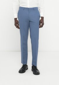 Άνδρας που φοράει ανοιχτό μπλε παντελόνι ραμμένο καλαίσθητα, λευκό πουκάμισο με κουμπιά και μαύρα δερμάτινα παπούτσια, στέκεται μπροστά σε λευκό φόντο.