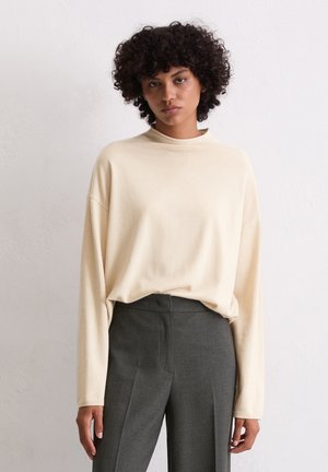 Beige Langarmoberteil mit hohem Kragen, lockerer Passform und weicher Textur, kombiniert mit dunkelgrauen maßgeschneiderten Hosen mit glatter Oberfläche.