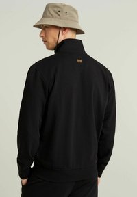 Sudadera negra con un alto cuello y puños de canalé. La parte posterior presenta una pequeña etiqueta rectangular en marrón y beige.