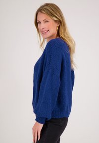 Blonde vrouw in een blauwe, gestructureerde trui en zwarte broek lacht en poseert opzij tegen een effen lichte achtergrond.