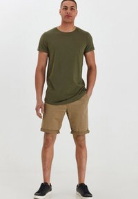 Olivgrünes Baumwoll-T-Shirt mit kurzen Ärmeln, kombiniert mit beigefarbenen, hochgerollten Shorts, die einfarbig sind und mit schwarzen Sneakers getragen werden.