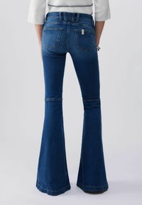 Femme portant un jean évasé taille haute bleu foncé avec des poches arrière et un haut blanc avec des coutures bleu marine.