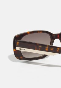 Ochelari de soare tortoiseshell maronii cu un cadru dreptunghiular gros, lentile nuanțate și un accent cu dungi aurii, având brandingul „QUAY” pe braț.