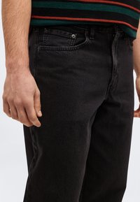 Χέρι άνδρα που ακουμπά δίπλα στην τσέπη μαύρου τζιν, φορόντας πράσινο και κόκκινο ριγέ πουκάμισο μέσα στην μέση.