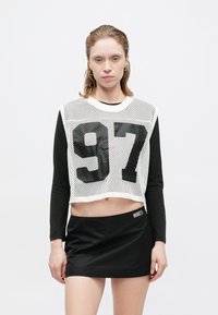 Geknipt langemouwen top in zwart-wit gaas met een groot zwart nummer "97." Gecombineerd met een strakke zwarte minirok met logo op de tailleband.