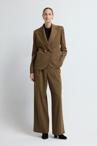 Brauner maßgeschneiderter Blazer mit zwei Fronttaschen und goldenen Knöpfen, kombiniert mit braunen, weit geschnittenen Hosen und schwarzen Stiefeletten.