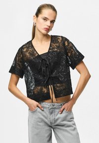 Črn čipkast kratki top s cvetličnim vzorcem, kratkimi rokavi, V-izrezom in detajlom sprednje zavezave; kombiniran s svetlo sivimi kavbojkami.