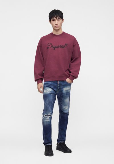 Νέος άνδρας φορώντας μωβ φούτερ Dsquared2, φθαρμένο μπλε τζιν και μαύρα αθλητικά παπούτσια, στέκεται μπροστά σε μια απλή φωτεινή επιφάνεια.