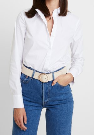 Femme portant une chemise blanche boutonnée rentrée dans un jean bleu avec une ceinture beige, une main posée dans la poche, sur un fond uni.