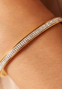 Pulsera de brazalete de oro que presenta una fila de cristales claros y brillantes engastados en una banda lisa. La pulsera resaltada sobre un fondo de piel.