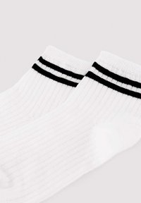 Calcetines tobilleros blancos con costillas y dos rayas negras cerca del puño, extendidos sobre una superficie clara.