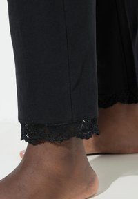 Zwarte katoenen broek met kanten rand aan de zoom. Heeft een gladde textuur en een aansluitend ontwerp, eindigend net boven de enkel, waarbij blote voeten zichtbaar zijn.