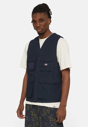 Chaleco utilitario de color azul marino, hecho de tela duradera, con múltiples bolsillos, un diseño con cuello en V y cierres de botones. Lleva puesto una camiseta ligera.