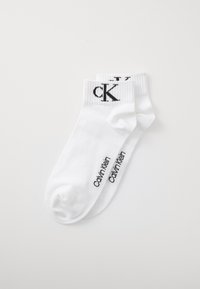 Un paio di calzini bianchi alla caviglia con il logo nero di Calvin Klein sul polsino e il nome del marchio stampato lungo il lato del piede, disposti piatti su uno sfondo bianco.