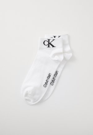 Promo de chaussettes blanches à chevilles avec le logo noir de Calvin Klein au niveau de la cheville et le nom de la marque imprimé sur le côté du pied, posées à plat sur un fond blanc.