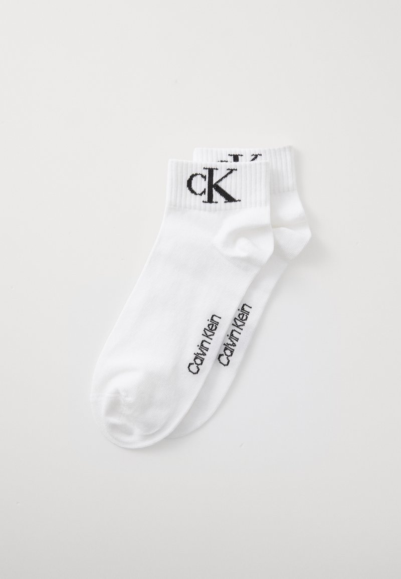 Par de meias brancas até ao tornozelo com o logótipo preto da Calvin Klein na margem e o nome da marca impresso ao longo do lado do pé, dispostas de forma plana sobre um fundo branco.